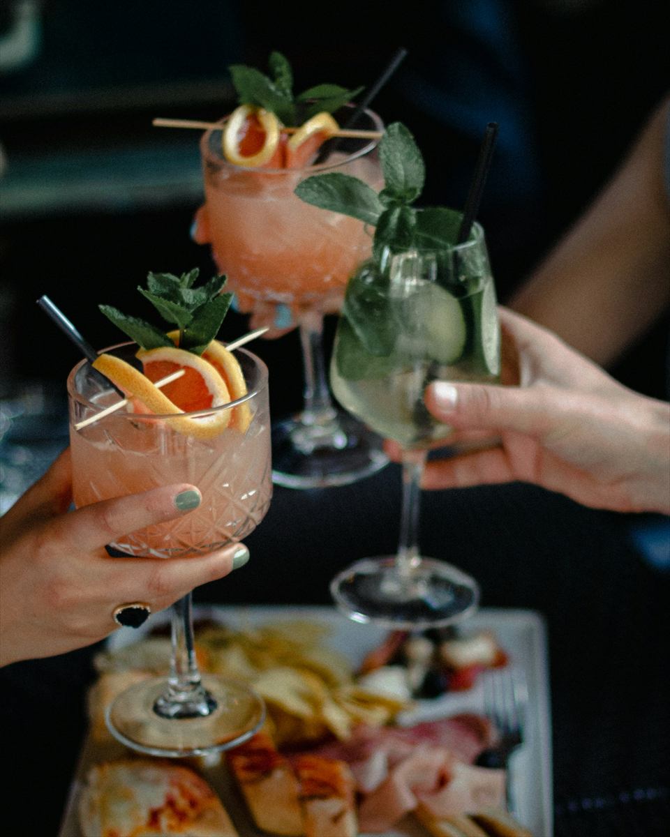 Mãos brindando com drinques coloridos sobre tábua de frios